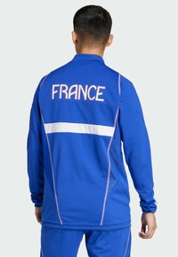 Veste de sport bleue avec "FRANCE" en lettres blanches au dos, accents roses et un détail à rayure horizontale blanche.