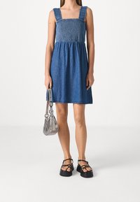 Vestido de mezclilla con cuerpo fruncido, escote cuadrado y tirantes anchos; combinado con un bolso plateado y sandalias de plataforma negras.