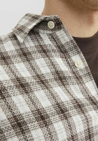 Skjorta med knappar i brun och vit rutigt mönster, tillverkad av texturerad tyg. Har en spetsig krage och synlig knappdetalj.