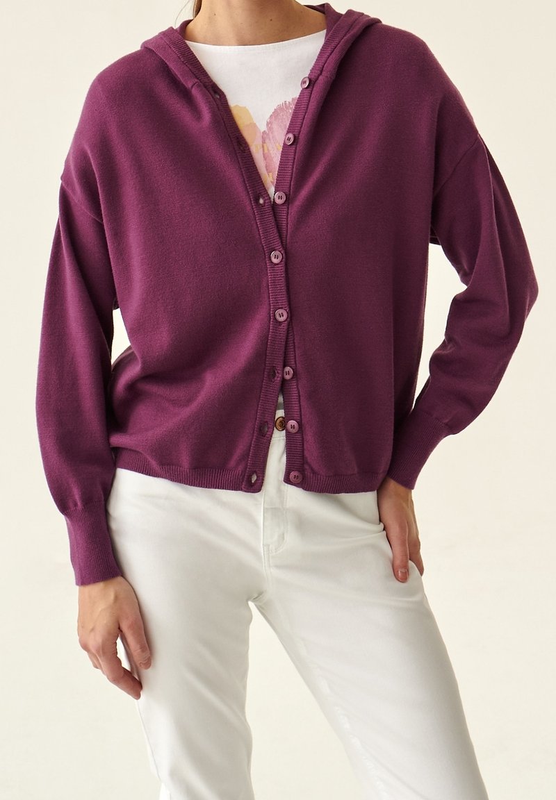 Femme portant un cardigan à capuche prune boutonné sur un t-shirt blanc avec motif et un pantalon blanc, debout avec une main sur la hanche.