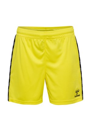 Ung person står iført sort sportst-shirt, lysegule shorts, sorte sokker og sorte sneakers mod en ensfarvet baggrund.