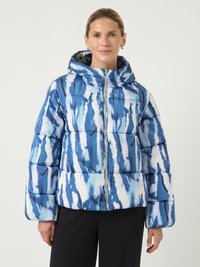 Moteris, dėvinti mėlynai-baltą abstrakčiai raštuotą pūkinę striukę su gobtuvu, stovi prieš vienodą šviesią fono.