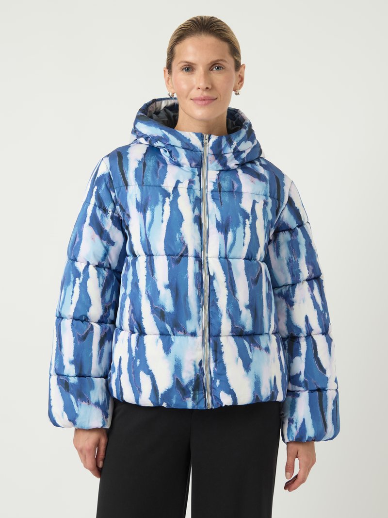 Moteris, dėvinti mėlynai-baltą abstrakčiai raštuotą pūkinę striukę su gobtuvu, stovi prieš vienodą šviesią fono.