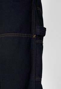 Pantalones de trabajo de mezclilla oscura con costuras amarillas en contraste, un bolsillo lateral y una tela resistente con un acabado texturizado.