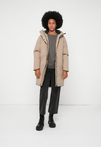 Mulher com cabelo afro, usando um casaco longo acolchoado bege, um suéter cinza, calças pretas cropped e botas pretas com atacadores, de pé contra um fundo branco.