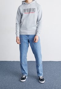 Felpa grigia con stampa del logo e tasca frontale, abbinata a jeans in denim azzurro chiaro con dettagli di cucitura e sneakers nere a bassa cima.