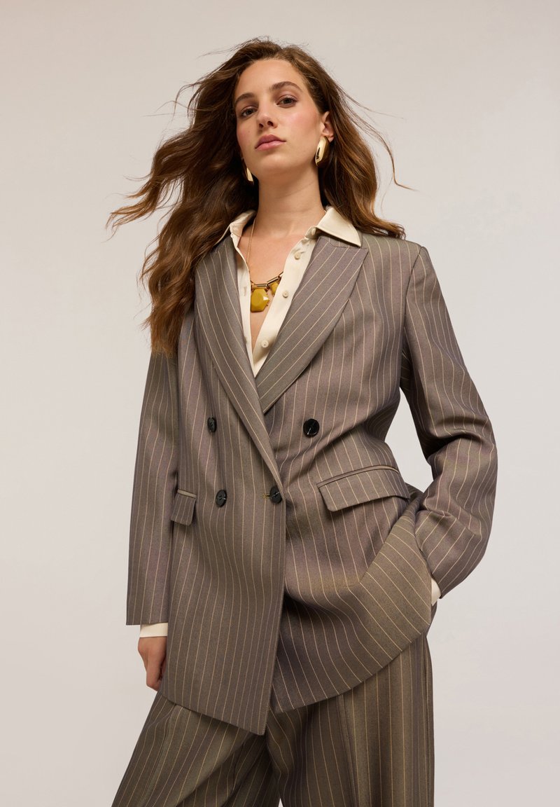 Blazer doppiopetto in tessuto grigio a righe con bottoni neri, con colletto a risvolto e due tasche frontali, abbinato a una camicia color crema.