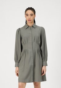 Rochie din oliva cu mânecă lungă și guler, cu pliseuri și închidere cu nasturi, având o textură netedă și talie ajustată.