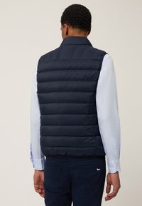 Mornarski pufasti prsluk s kvadratnim dizajnom, visokim okovratnikom i glatkom teksturom. Nosi se preko svijetlos plave košulje s dugim rukavima, koja ima detalje s gumbima.