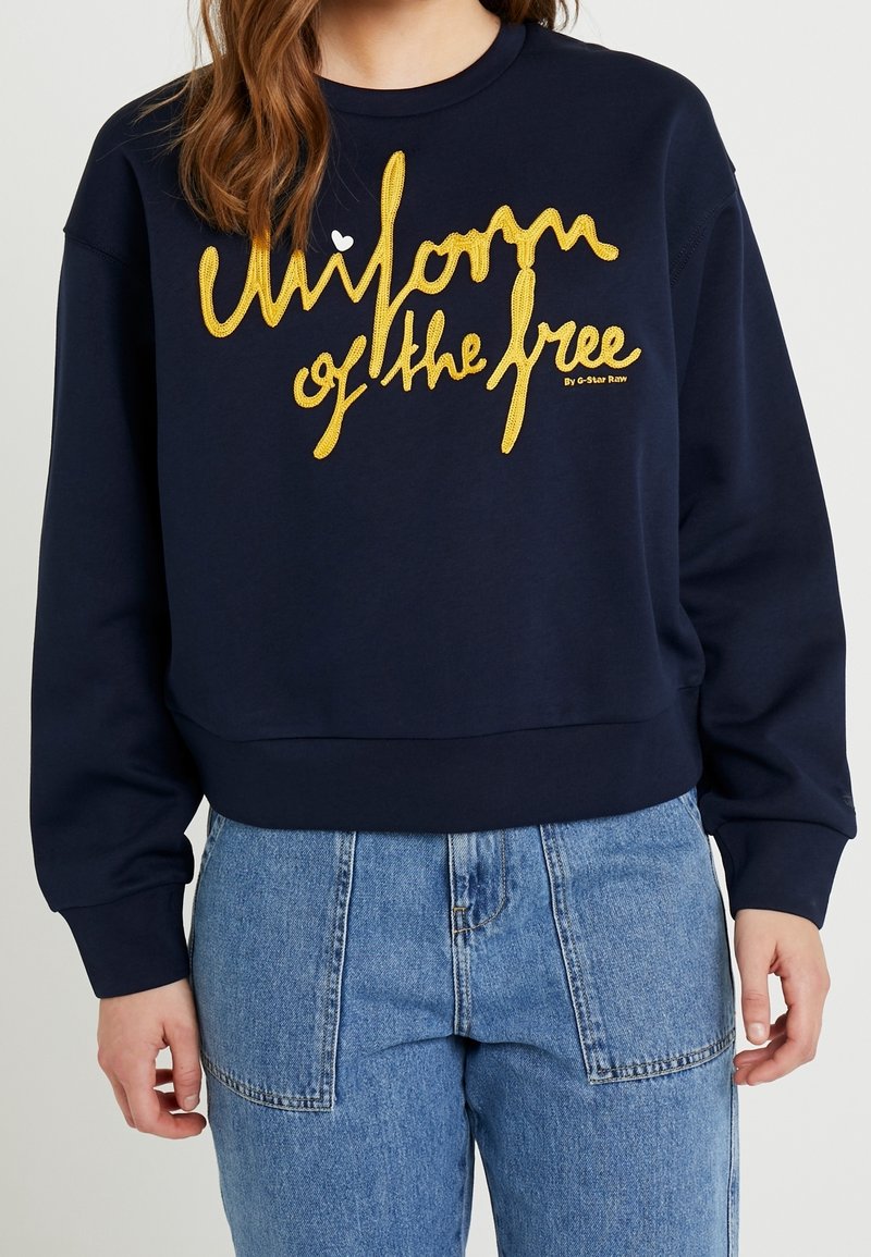Felpa blu navy con grande scritta gialla ricamata "uniforme dei liberi", con polsini e orlo a coste. Abbinata a jeans azzurri.