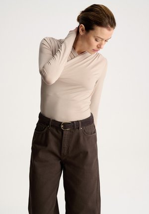 Kobieta w beżowej bluzce z długim rękawem i ciemnobrązowych spodniach dotykająca szyi, patrząca w dół na jednolitym jasnym tle.