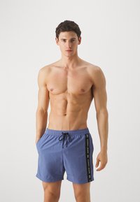 Pantaloni da bagno blu realizzati in tessuto leggero, con una cintura elastica dotata di cordino e un logo decorativo laterale.