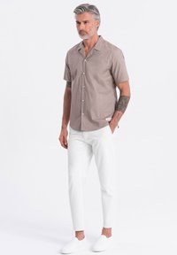 Kurzärmliges, hellbraunes Hemd mit Knopfleiste und Kragen, kombiniert mit weißen, schmal geschnittenen Hosen und weißen Slippern, die ein minimalistisches Design aufweisen.
