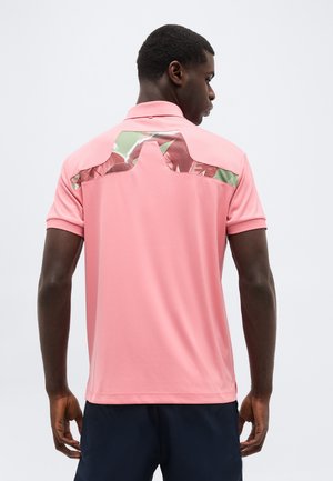 Man wearing a pink short-sleeve polo shirt with reflective silver and green detail across the upper back, paired with dark shorts.