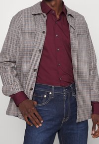Kurtka w kratę w odcieniach brązu i beżu z zapinanym przodem, zestawiona z bordową koszulą i ciemnoniebieskimi dżinsami. Gładka faktura i dopasowany krój.