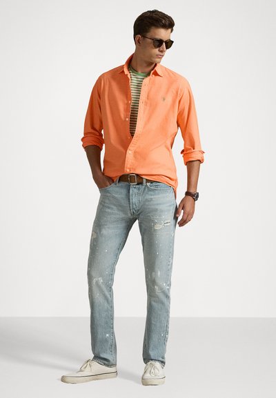 Oranssi napillinen paita raidallisen t-paidan päällä, yhdistettynä haalistuneisiin sinisiin kulutettuihin farkkuihin ja valkoisiin lenkkareihin. Asusteina aurinkolasit ja kello.