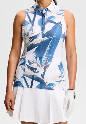 Frau trägt ärmelloses Golfshirt mit Kragen, blau-beigem Blumenmuster und weißem plissiertem Rock, hält weißen Golfhandschuh an der rechten Hand.