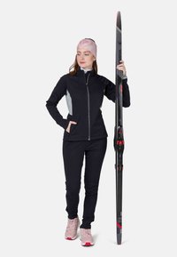 Sort træningsjakke med grå detaljer, sorte bukser og lyserøde træningssko. Model holder en sort og rød ski. Glat stof, tætsiddende design.