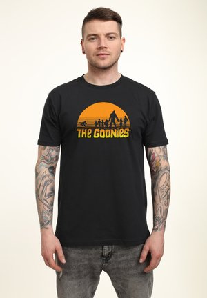 Sort t-shirt med et levende orange solnedgangsdesign med silhuetter og teksten "The Goonies" i gult nederst.