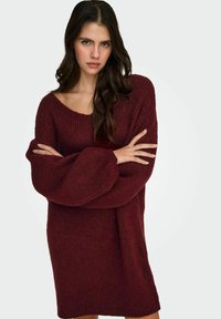 Sweter dziergany w ciemnym bordowym kolorze o luźnym kroju, szerokich rękawach i dekolcie w kształcie litery U. Tekstura wydaje się miękka i przytulna, odpowiednia na chłodniejszą pogodę.