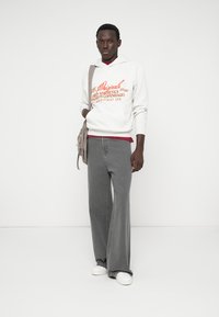 Jeans largas cinzentas combinadas com um hoodie cinzento claro com texto gráfico em vermelho. Uma bolsa de ombro bege complementa o look.