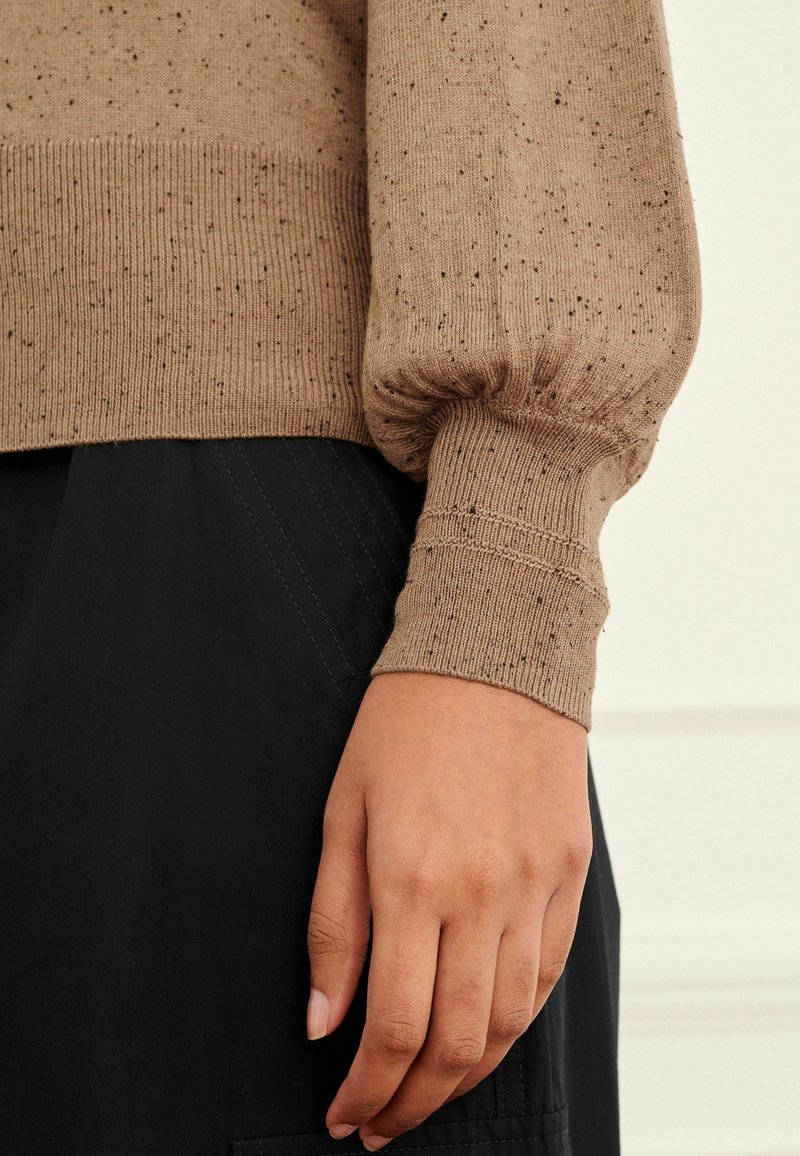 Pull en maille brun, tacheté, avec des poignets côtelés, présenté avec un vêtement noir. Les poignets sont volumineux, avec une finition texturée, mettant en valeur le design du tissu.