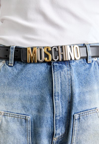 Fekete bőrből készült öv, nagy arany és ezüst "MOSCHINO" felirattal; világoskék farmernadrággal párosítva, amelyen jól látható varrási részletek vannak.