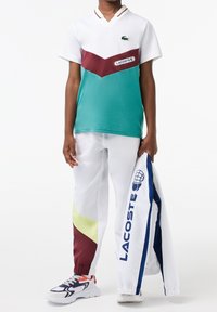Zielono-biała siateczkowa koszulka polo z bordowymi akcentami, w połączeniu z białymi spodniami z kolorowymi panelami bocznymi oraz kurtką Lacoste.