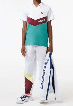 Zielono-biała siateczkowa koszulka polo z bordowymi akcentami, w połączeniu z białymi spodniami z kolorowymi panelami bocznymi oraz kurtką Lacoste.