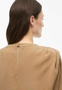 Blouse marron avec un col rond, tissu doux, léger brillant, et fermeture à boutons dans le dos ; présente des détails plissés aux épaules.