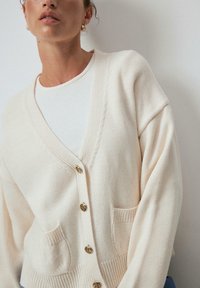 Cardigan color crema con scollo a V, cinque bottoni dorati, tasche anteriori e polsini a coste. Texture in maglia morbida con una vestibilità rilassata.
