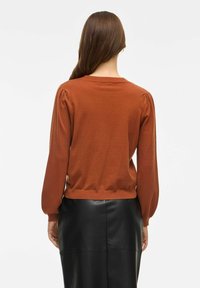 Pull couleur rouille avec des manches bouffantes et un col rond, associé à une jupe en cuir noire, mettant en valeur un design confortable et ajusté.