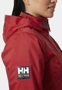 Czerwona wodoodporna kurtka z teksturowanym wykończeniem, z ściąganym kapturem, czarną łatką z logo i wysokim kołnierzem.