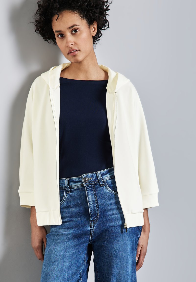 Light yellow zip-up hoodie with a spacious hood, worn over a navy top. Paired with high-waisted blue denim jeans, featuring a button closure.
