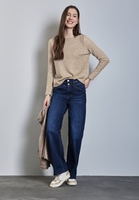 Top beige a maniche lunghe in tessuto testurizzato, abbinato a jeans a gamba larga blu scuro. La modella tiene una giacca marrone chiaro e indossa sneakers beige.