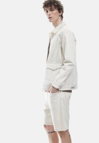 Chaqueta de denim blanca con dos bolsillos frontales y detalles de botones, combinada con pantalones cortos a juego. Textura suave, diseño limpio, estilo minimalista.