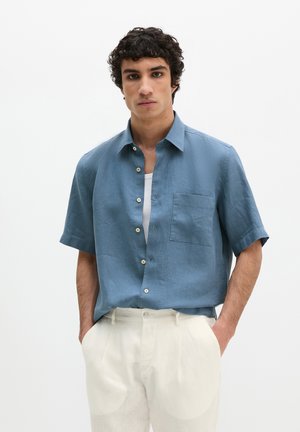 Mann mit lockigem Haar, der ein blaues kurzärmeliges Hemd über einem weißen Hemd und weiße Hosen trägt, mit den Händen in den Taschen stehend.