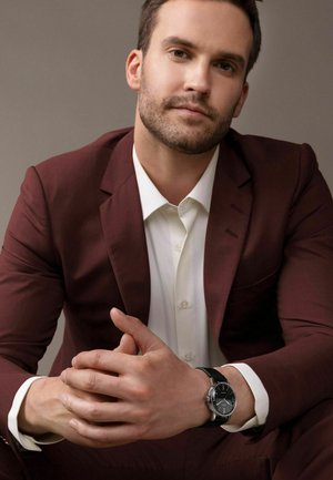 Montre-bracelet noire avec accents argentés, portée sur une main. Le sujet est en costume bordeaux et chemise blanche, assis avec les mains jointes.
