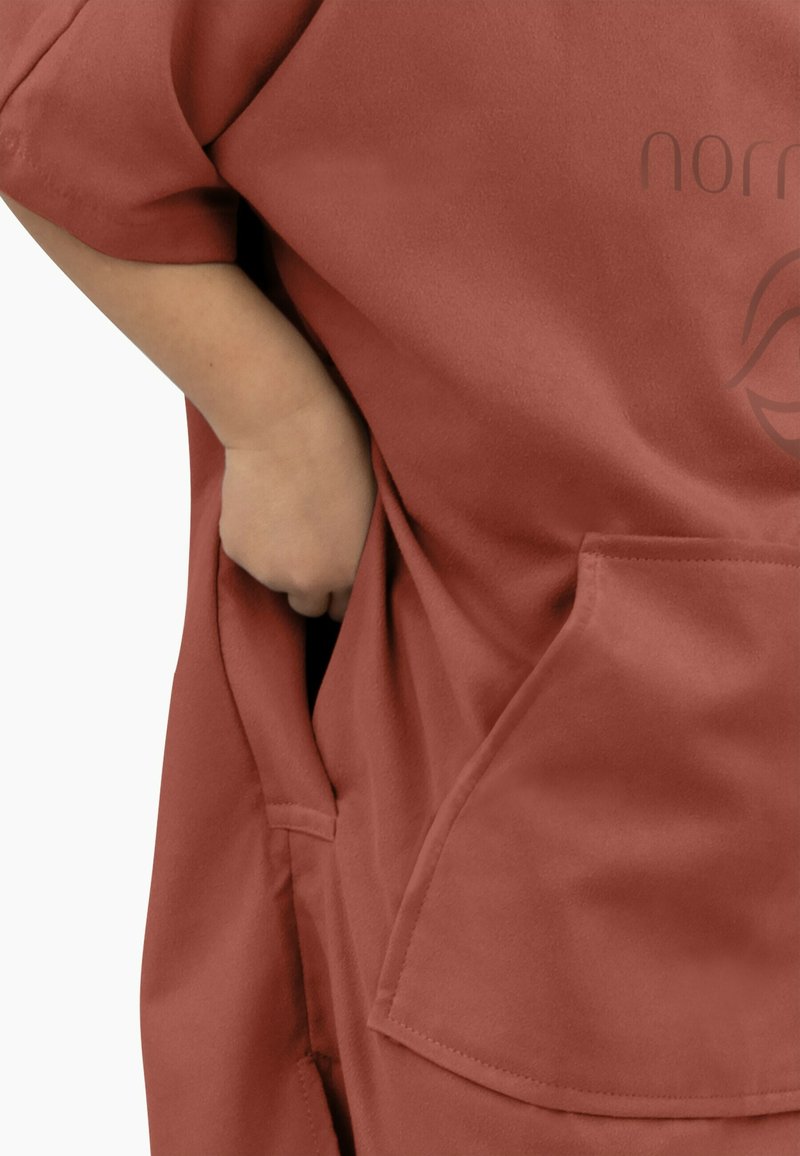 Kurzärmliges, rostfarbiges Kleidungsstück mit glatter Textur und vorderen Taschen. Verfügt über ein dezent auf dem Stoff gedrucktes Markenlogo.