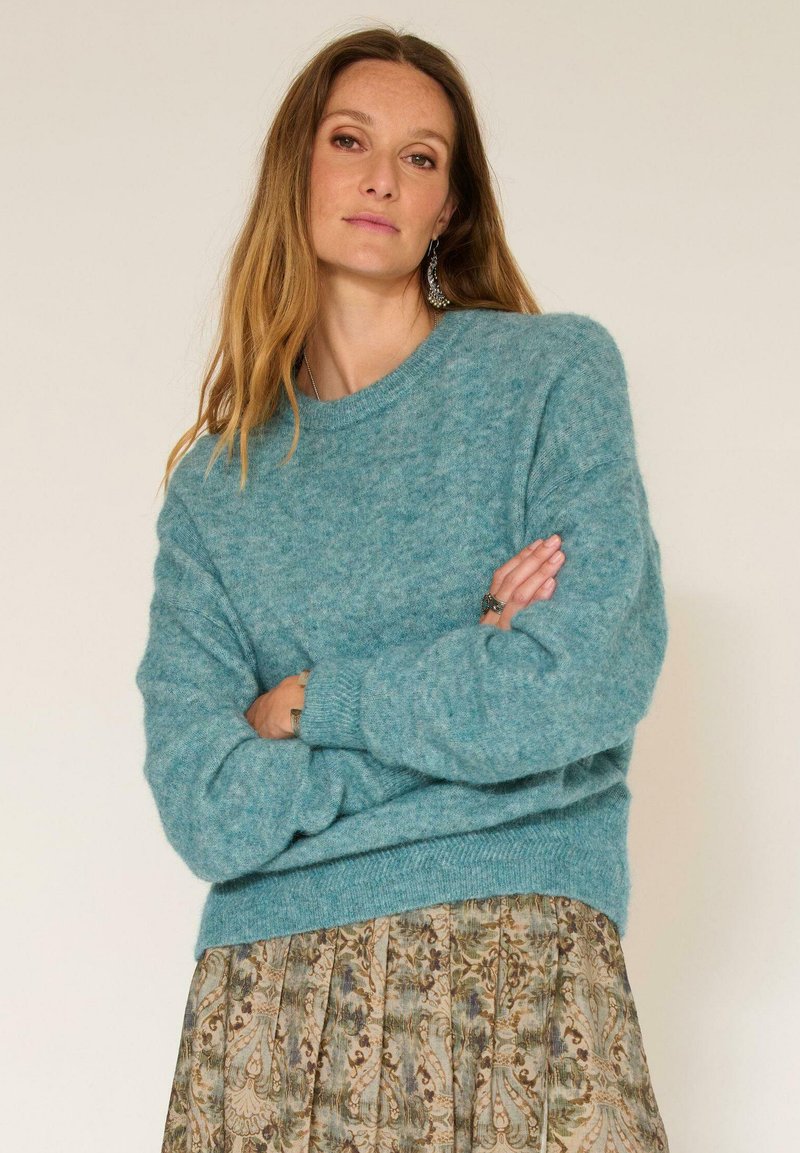 Pull en tricot turquoise avec un col rond, des épaules tombantes, des poignets côtelés et une coupe décontractée, associé à une jupe à motifs.
