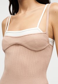 Εγγύς λήψη ενός ατόμου που φοράει ένα ριμπ πλεκτό φόρεμα καρμί σε ανοιχτό ροζ χρώμα, φορεμένο πάνω από ένα λευκό αμάνικο τοπ με λεπτούς ιμάντες.