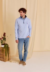 Lichtblauw, getextureerd sweater met een ritssluiting bij de kraag en logo op de borst, gecombineerd met lichte jeans en beige schoenen, tegen een neutrale achtergrond.