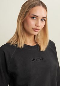 Giovane donna con capelli biondi lisci, che indossa una felpa nera con la scritta "freddy" ricamata sul petto, sfondo beige neutro.
