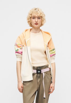 Άτομο με ξανθά, σγουρά μαλλιά που φοράει φούτερ με φερμουάρ σε ροδακινί και εκρού χρώμα, μπεζ μπλούζα, καφέ παντελόνι και ανοιχτοροζ ζώνη, στέκεται μπροστά σε απλό φόντο.