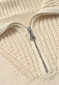 Gestrickter, hellbeiger Pullover mit einem Frontreißverschluss, der über einen strukturierten Stoff und einen gerippten Kragen verfügt. Silberne Reißverschlussbeschläge sorgen für das gewisse Etwas.