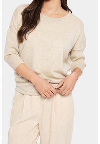 Beige Strickpullover mit entspannter Passform, weitem rundem Ausschnitt und gerippten Details an den 3/4-Ärmeln. Kombiniert mit hellbeigen Hosen.