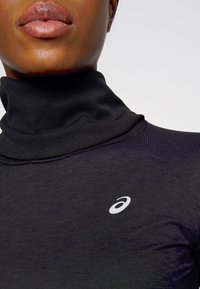 Maglione a dolcevita nero con una texture a coste, caratterizzato da un piccolo logo bianco sul lato sinistro. La vista ravvicinata evidenzia i dettagli del tessuto.