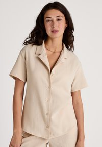 Kortebloes, met kraag, in lichtbeige stof, met een knoopsluiting aan de voorkant, subtiele textuur en een losse pasvorm.