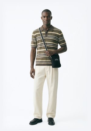 Homme portant un polo à manches courtes rayé marron, un pantalon crème, des mocassins noirs et un sac bandoulière noir, debout devant un fond blanc.