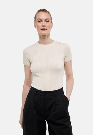 Femme aux cheveux blonds attachés en chignon bas, portant un haut beige côtelé à manches courtes et un pantalon noir taille haute, debout avec une main dans la poche.